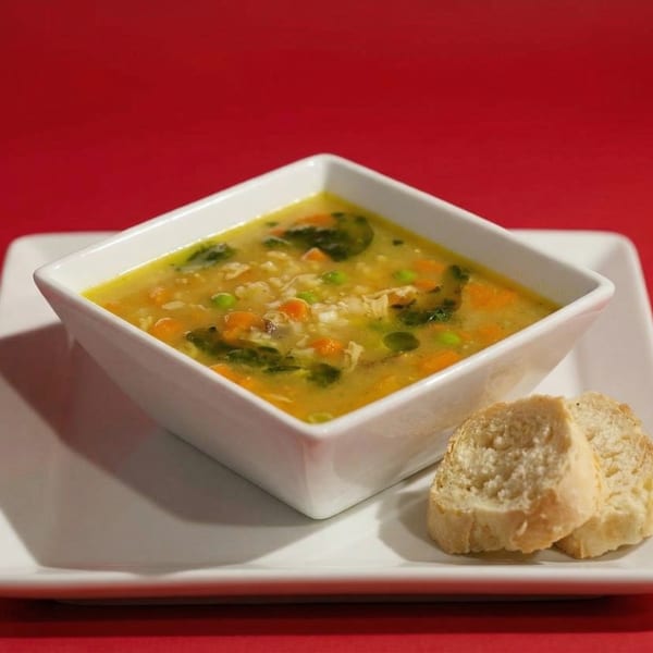 Hearty chicken and vegetable soup with two slices of bread on a white plate against a red background.